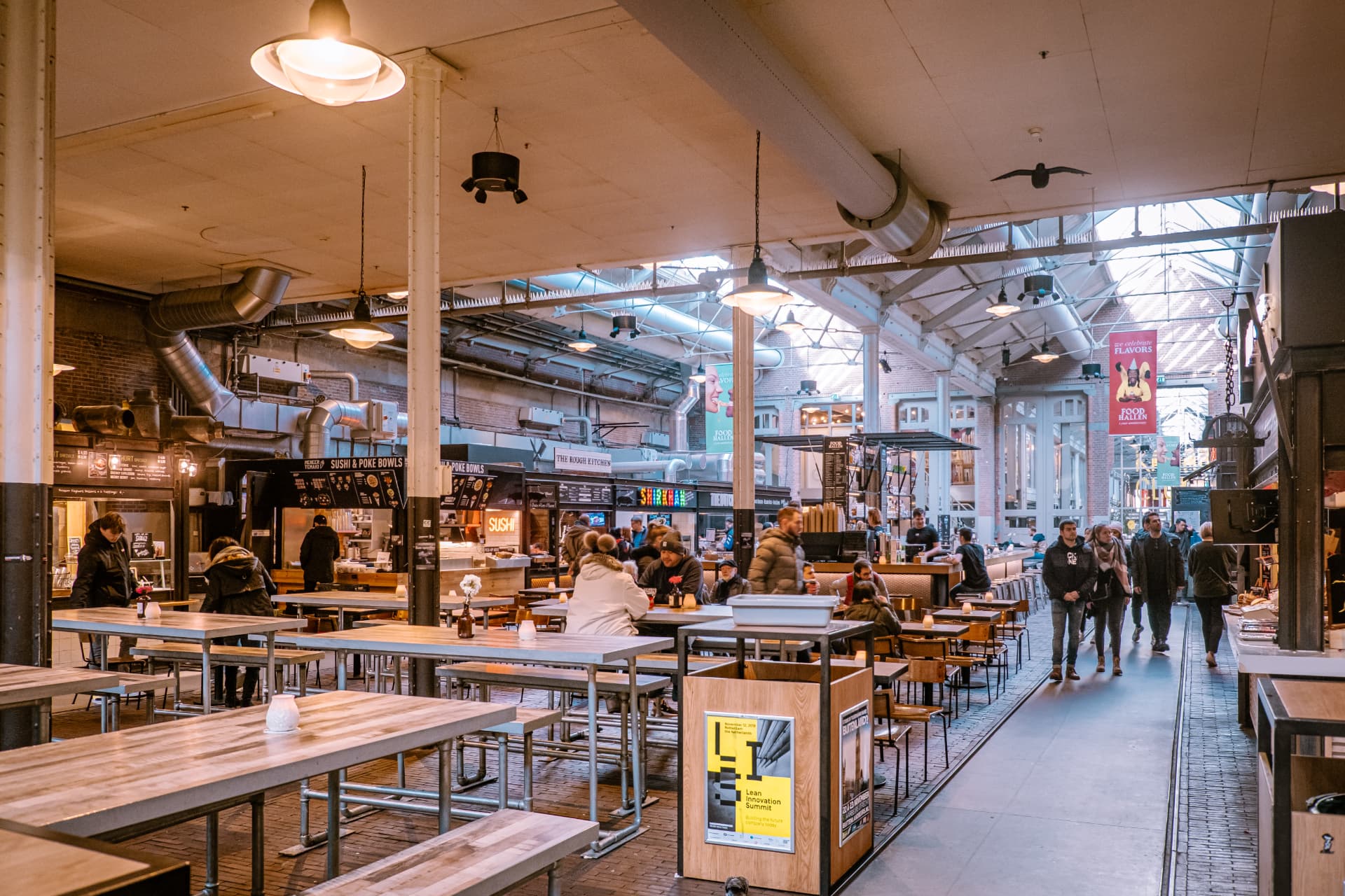 Long wooden tables, hanging lamps and food stalls under the iron and glass roof of Foodhallen in De Hallen, Amsterdam