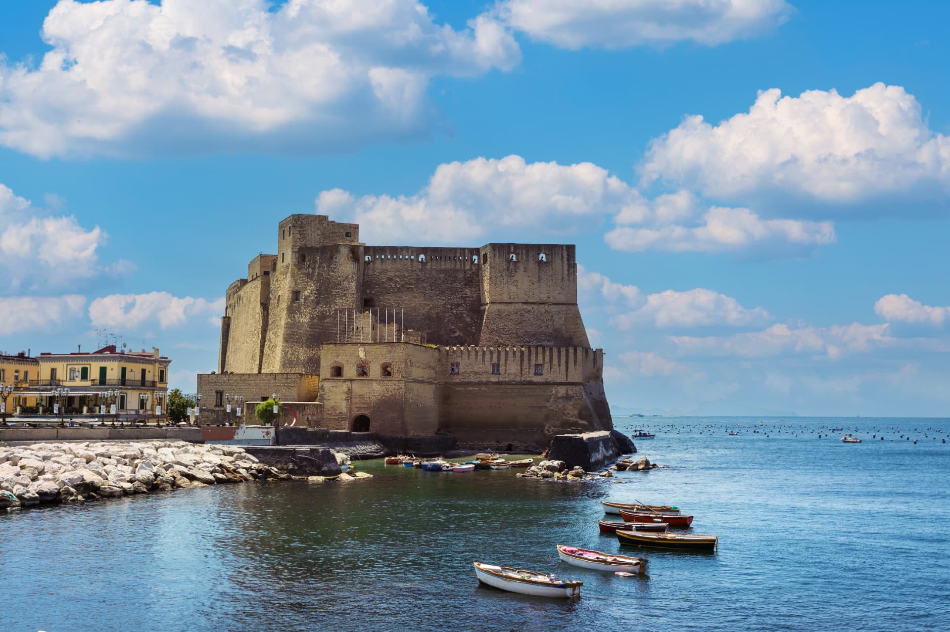Castel dell'Ovo fortress on the Naples waterfront under a bright blue sky with small boats moored around the jetty