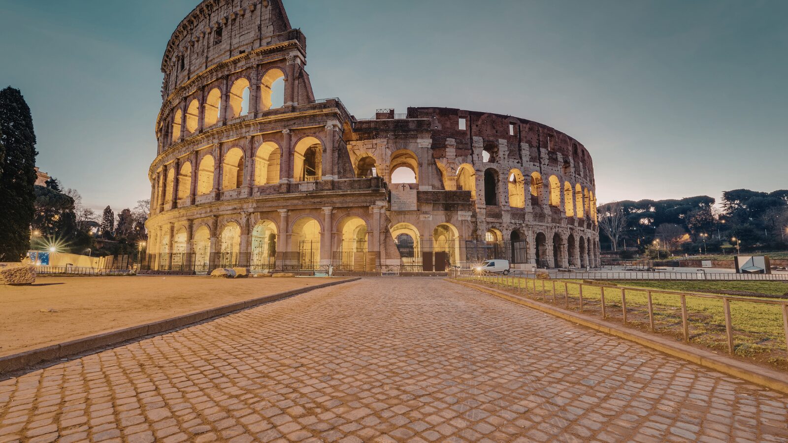 Colosseum illuminated at twilight from cobblestone road in Rome 2026 — golden lights highlighting the ancient arches against a blue evening sky