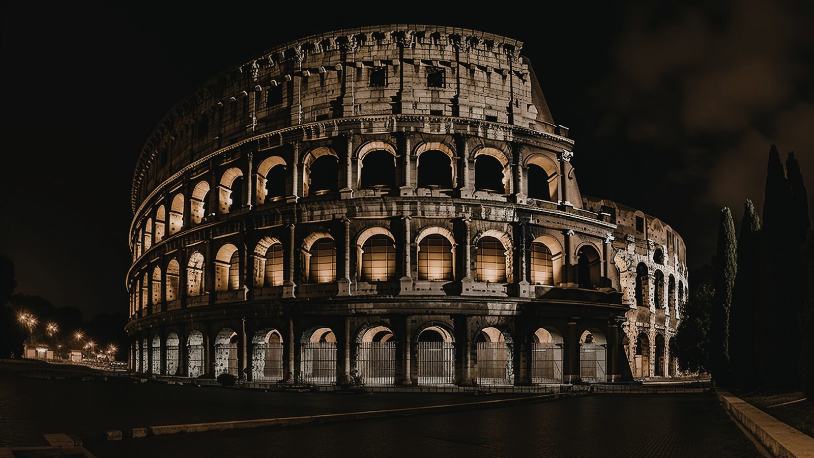 Colosseum illuminated at night in Rome 2026 — dramatic golden lighting reveals three tiers of arches against the dark sky