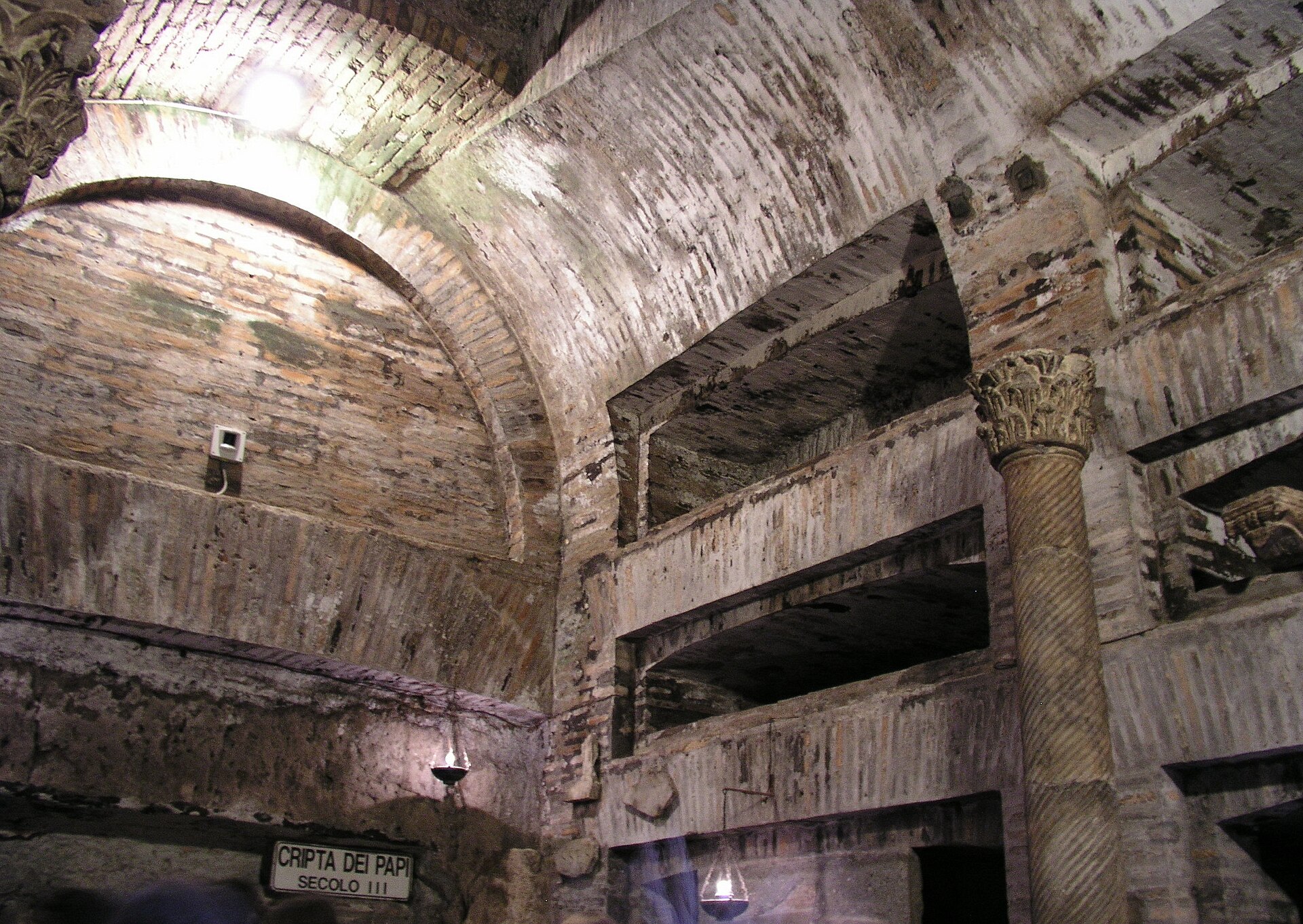 The Crypt of the Popes in the Catacombs of San Callisto, a vaulted tufa chamber with stacked burial niches, an ancient spiral column and a sign reading Cripta dei Papi Secolo III