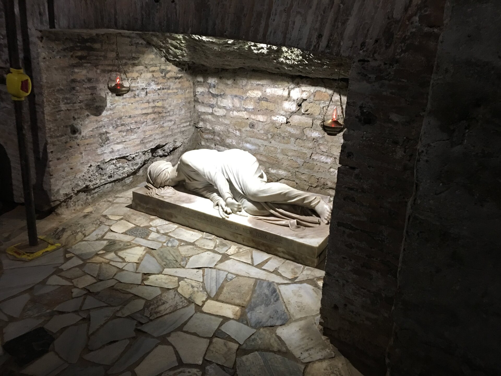 Marble statue of Saint Cecilia lying on her side in the crypt of Saint Cecilia in the Catacombs of San Callisto, lit by two hanging oil lamps