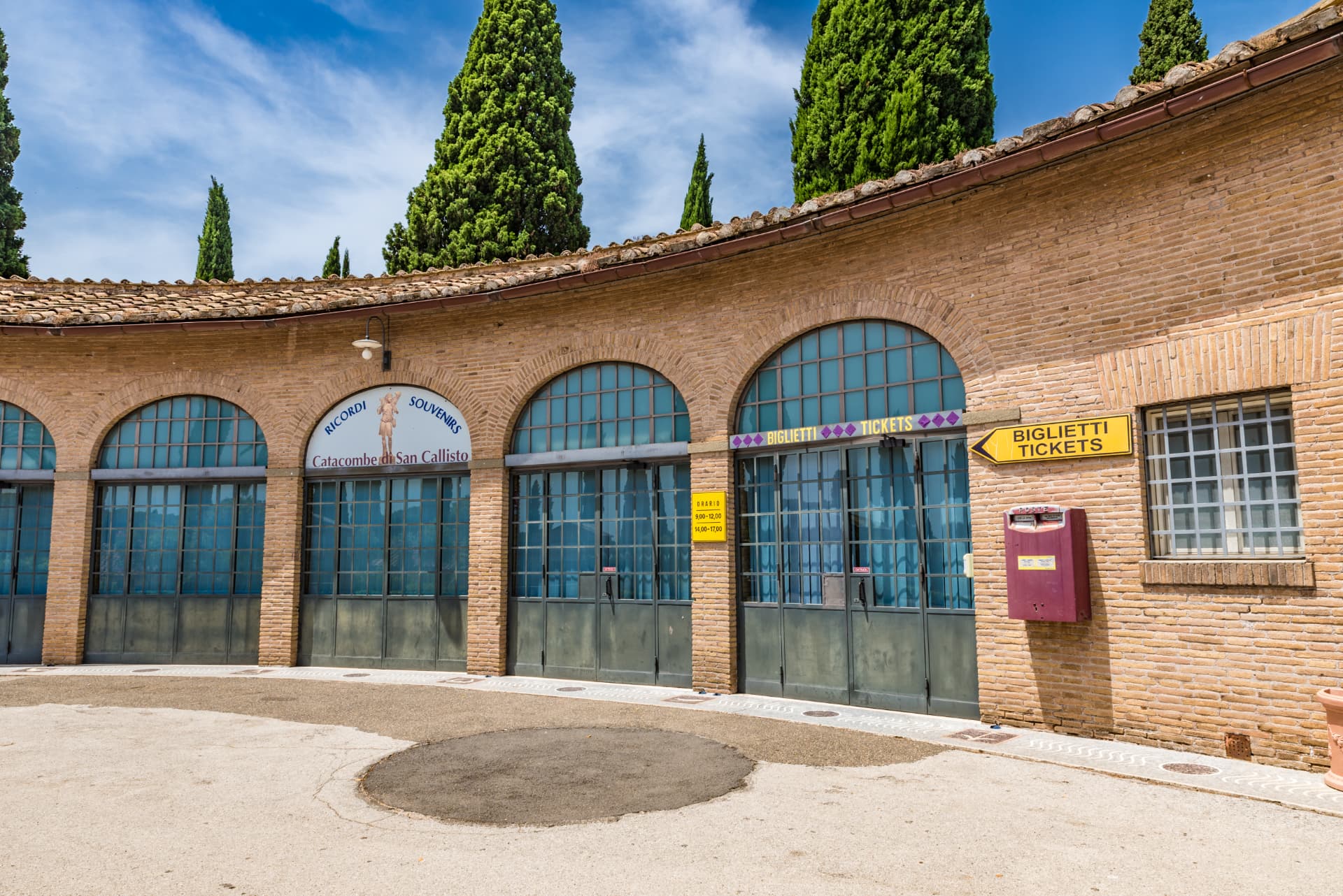 Ticket office and souvenir arcade at the Catacombe di San Callisto on the Appian Way, with cypress trees above the brick building