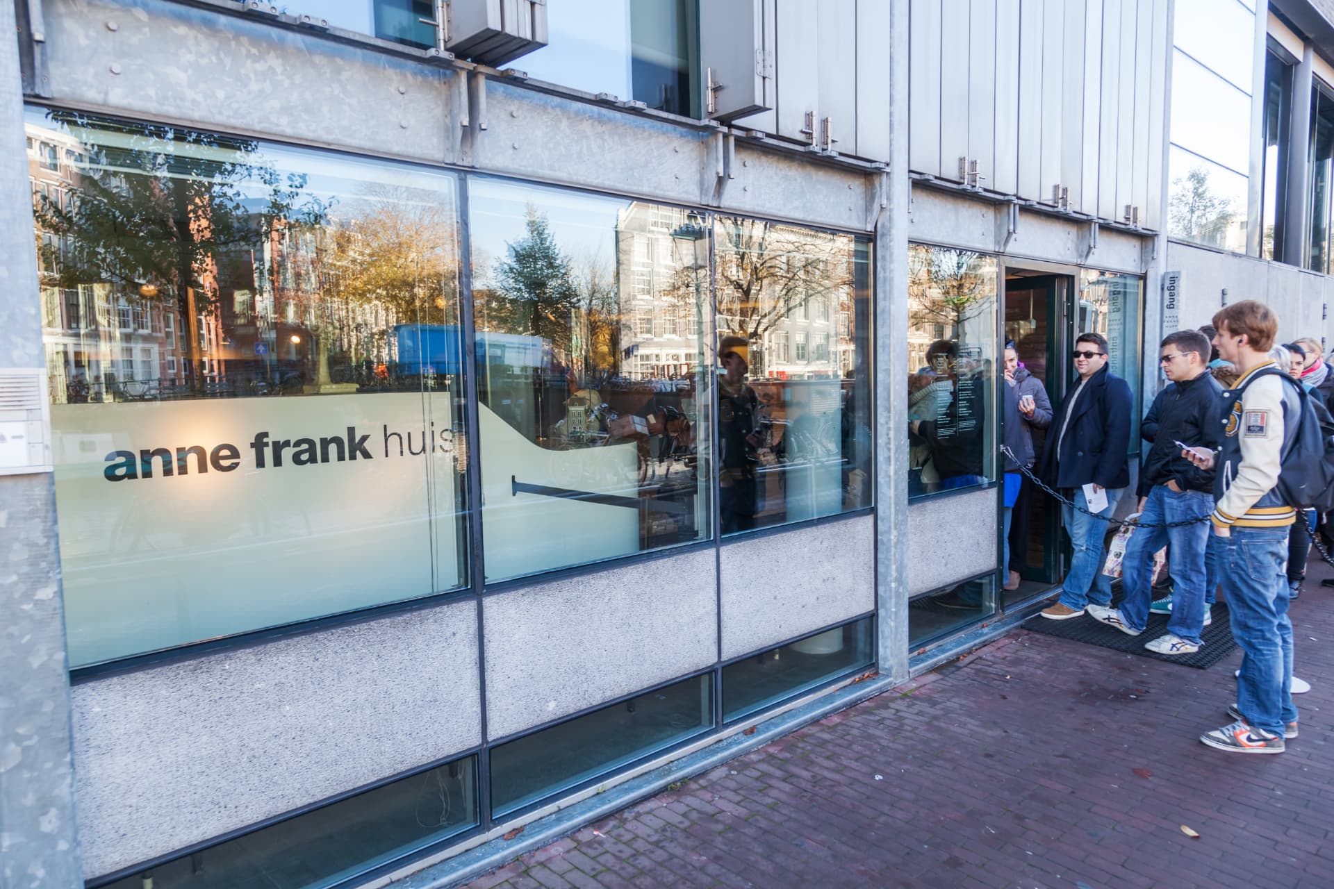 Visitors waiting at the modern glass museum entrance of the Anne Frank House, with the anne frank huis lettering on the frosted glass