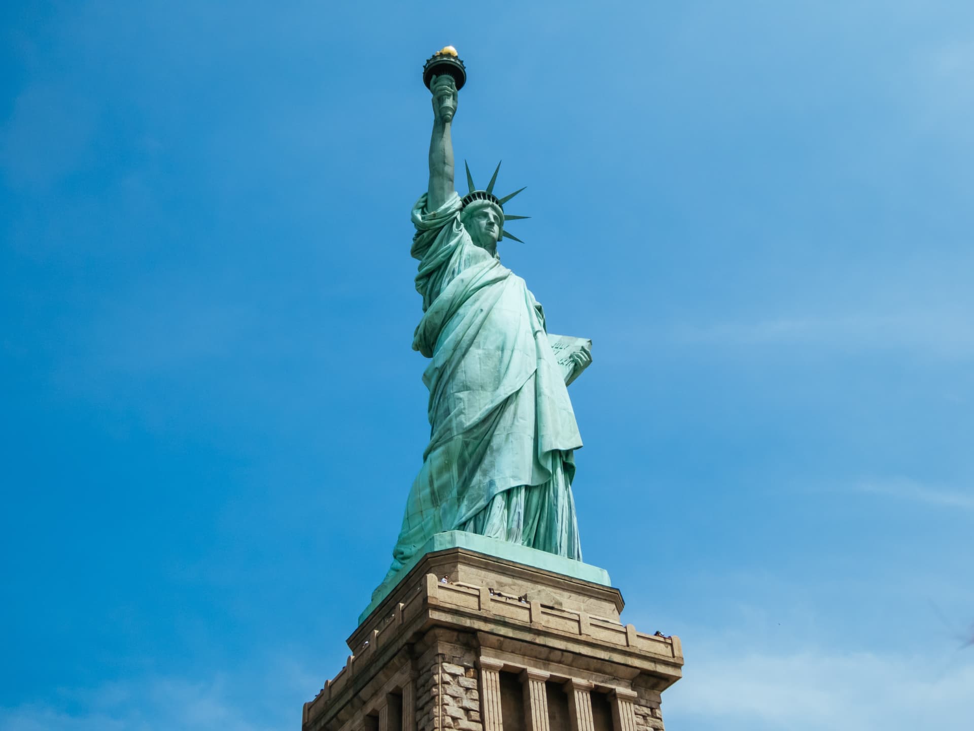 The Statue of Liberty photographed from below against a blue sky, showing the copper-green robes, tablet and raised torch