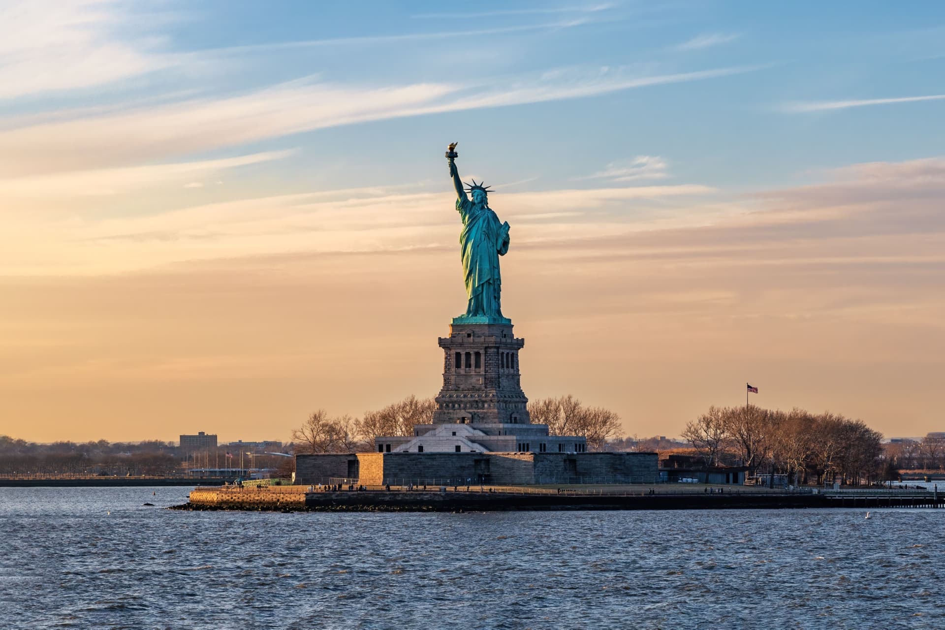 The Statue of Liberty silhouetted against a golden sunset sky, seen across the water from the harbour