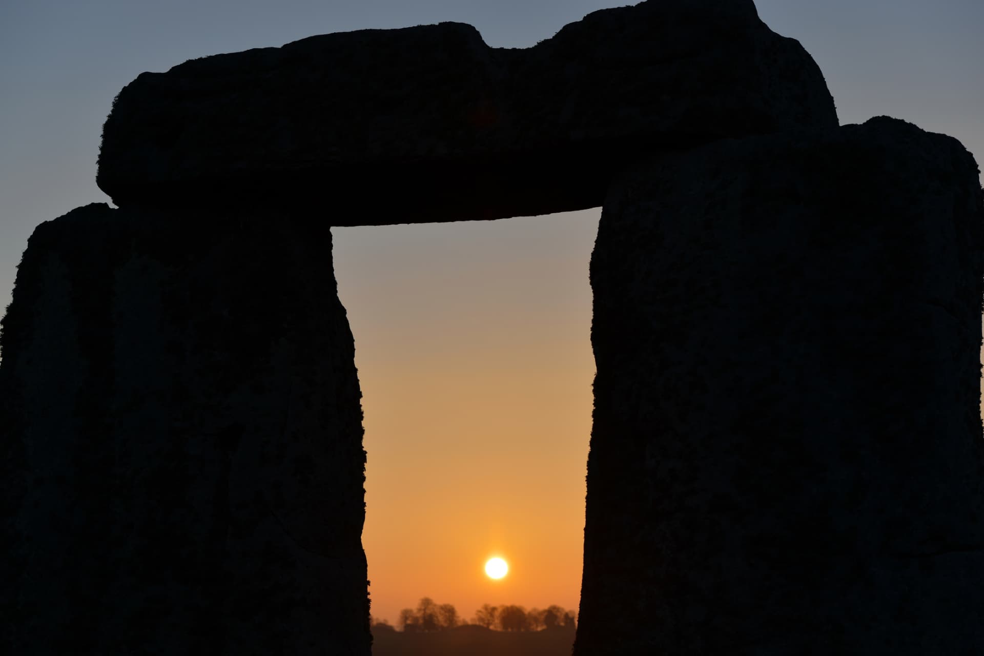 The sun setting between the uprights of a Stonehenge trilithon, the stones silhouetted black against an orange sky